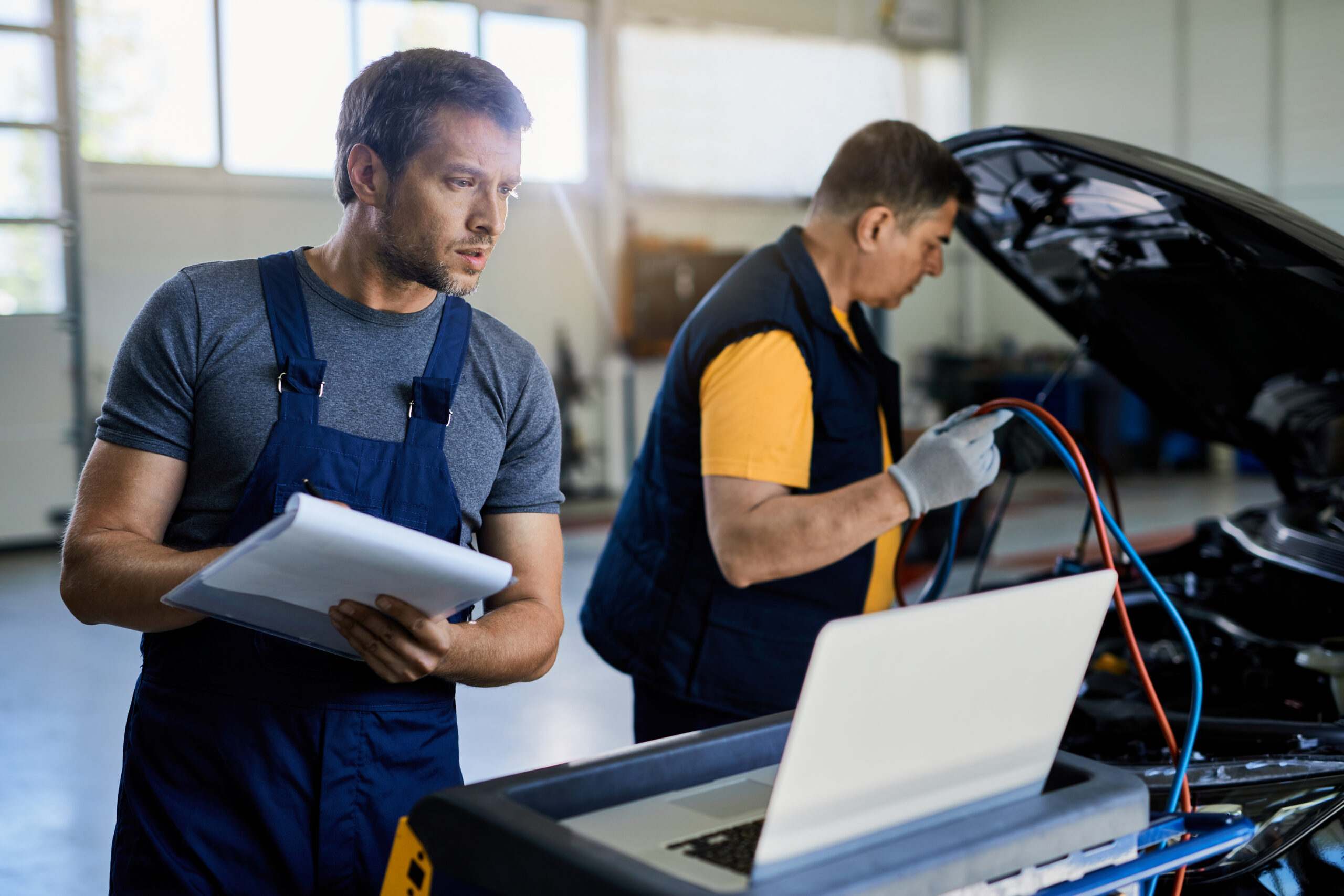 Auto repairman taking notes while using laptop in a workshop.